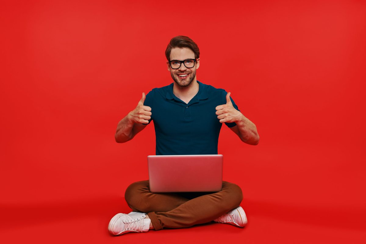 Happy young man in casual wear carrying computer and showing his thumbs up while sitting against red background